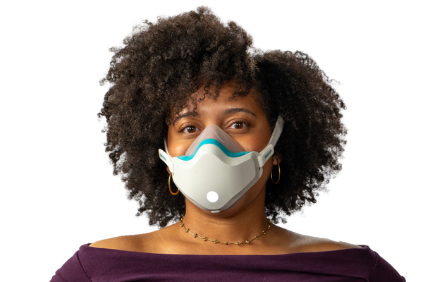 A woman with curly dark hair wearing a purple off-the-shoulder long-sleeve top, blue jeans, and a white face mask, standing with hands on hips against a solid white background.