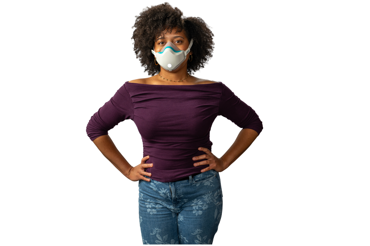 A woman with curly dark hair wearing a purple off-the-shoulder long-sleeve top, blue jeans, and a white face mask, standing with hands on hips against a solid white background.